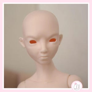 May include: A pale pink doll head with orange eye sockets. The doll has a smooth, blank face with a slightly furrowed brow and a small, pursed mouth. The head is bald and has small ears. The doll's neck and shoulders are also visible.