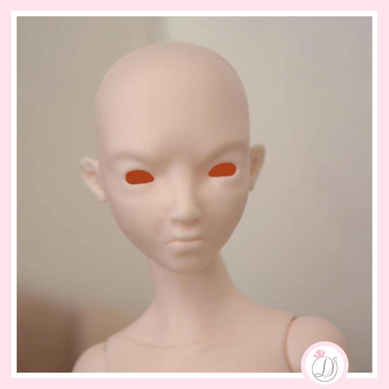 May include: A pale pink doll head with orange eye sockets. The doll has a smooth, blank face with a slightly furrowed brow and a small, pursed mouth. The head is bald and has small ears. The doll's neck and shoulders are also visible.