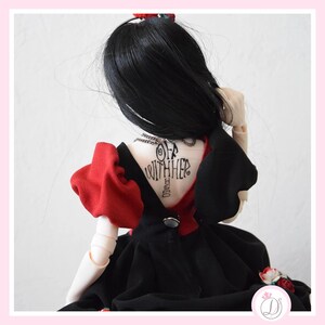 May include: A doll wearing a black dress with red accents and a tattoo that says "Off With Her". The doll has black hair and is posed from the back. The dress has a button closure and a small floral detail.