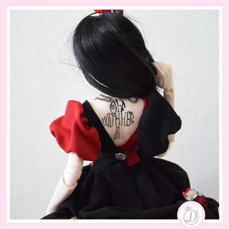 May include: A doll wearing a black dress with red accents and a tattoo that says "Off With Her". The doll has black hair and is posed from the back. The dress has a button closure and a small floral detail.
