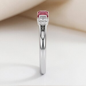 May include: A silver-toned ring featuring a rectangular-cut deep red gemstone at the center, flanked by a cluster of three small, round, clear stones on each side, set in a polished metal band with a smooth, reflective finish and tapered design.