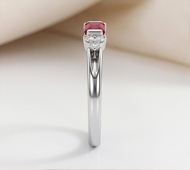 May include: A silver-toned ring featuring a rectangular-cut deep red gemstone at the center, flanked by a cluster of three small, round, clear stones on each side, set in a polished metal band with a smooth, reflective finish and tapered design.