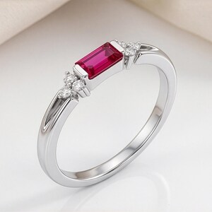 May include: A silver-toned ring featuring a central rectangular baguette-cut red gemstone. The band is accented on both sides of the center stone by a cluster of three small, round, clear, sparkling stones set into the metal shank.