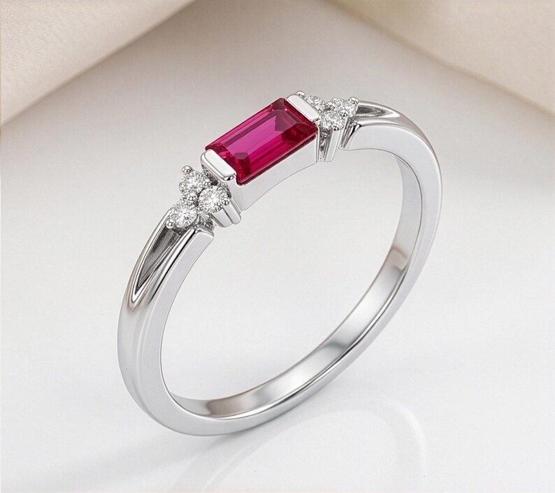 May include: A silver-toned ring featuring a central rectangular baguette-cut red gemstone. The band is accented on both sides of the center stone by a cluster of three small, round, clear, sparkling stones set into the metal shank.