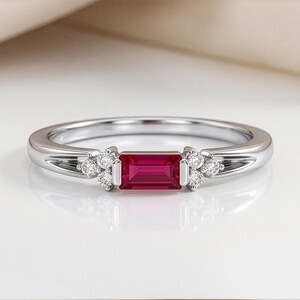 May include: A silver-toned ring featuring a central rectangular baguette-cut red gemstone. The stone is flanked on both sides by a cluster of three small, round, clear, brilliant-cut stones set into the polished metal band of the ring.
