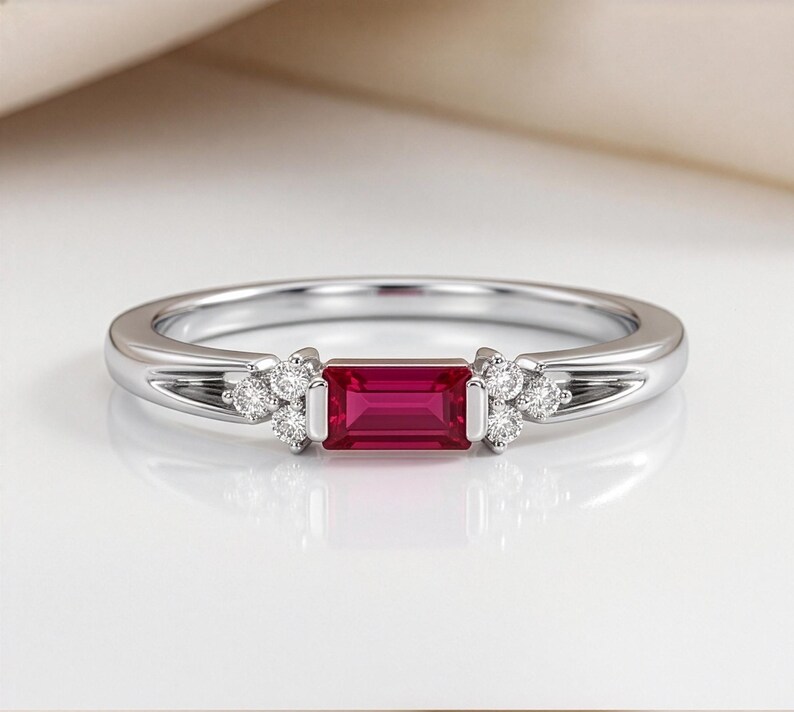 May include: A silver-toned ring featuring a central rectangular baguette-cut red gemstone. The stone is flanked on both sides by a cluster of three small, round, clear, brilliant-cut stones set into the polished metal band of the ring.