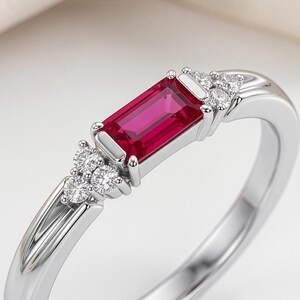 May include: A silver ring featuring a rectangular baguette-cut vibrant red gemstone set in the center. The stone is flanked on both sides by a cluster of three small, round, clear white accent stones set into the polished silver band.