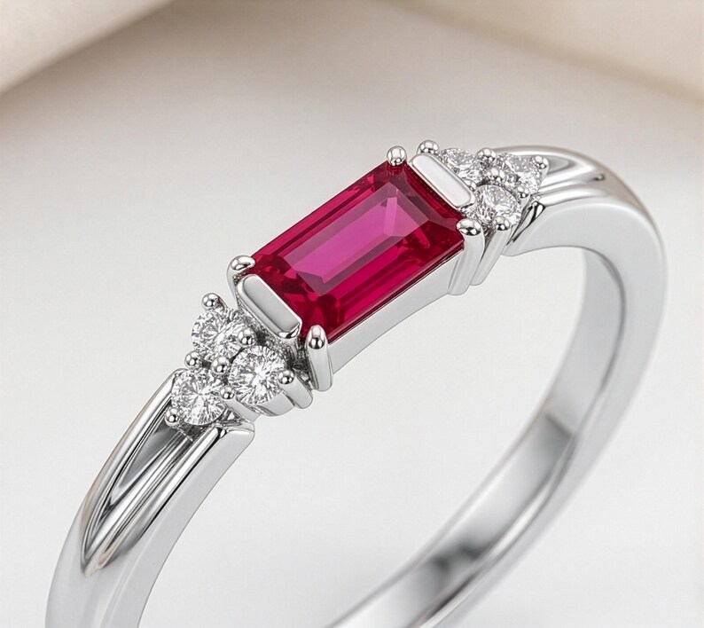 May include: A silver ring featuring a rectangular baguette-cut vibrant red gemstone set in the center. The stone is flanked on both sides by a cluster of three small, round, clear white accent stones set into the polished silver band.