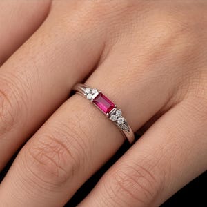 May include: A silver-toned ring featuring a central rectangular baguette-cut vibrant red gemstone. The stone is flanked on both sides by a cluster of three small, round, clear white stones set into the polished metal band of the ring.