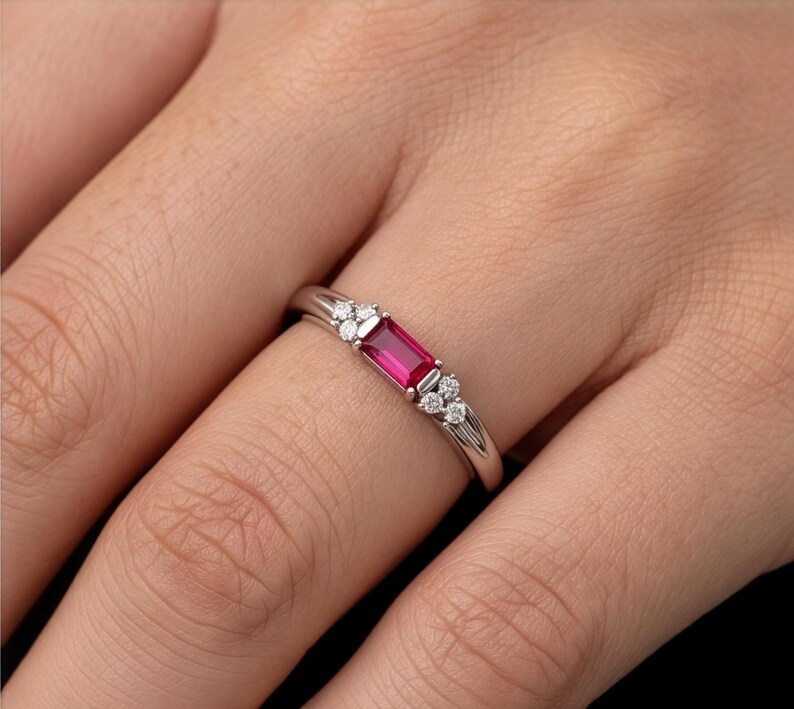 May include: A silver-toned ring featuring a central rectangular baguette-cut vibrant red gemstone. The stone is flanked on both sides by a cluster of three small, round, clear white stones set into the polished metal band of the ring.