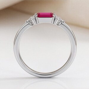 May include: A silver-toned ring featuring a rectangular, emerald-cut vibrant pink gemstone set horizontally at the center. The band is accented on both sides of the central stone by a cluster of three small, clear, round-cut sparkling stones.