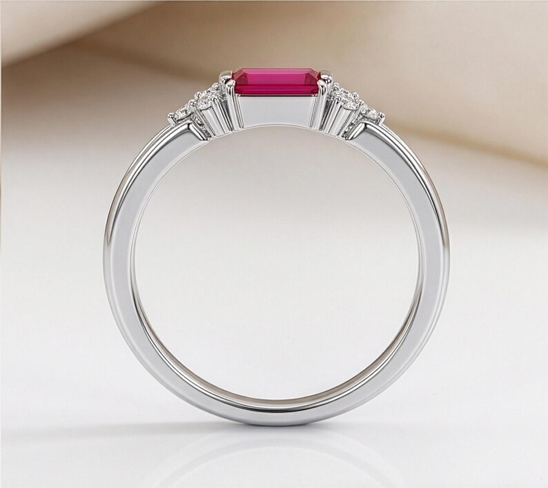 May include: A silver-toned ring featuring a rectangular, emerald-cut vibrant pink gemstone set horizontally at the center. The band is accented on both sides of the central stone by a cluster of three small, clear, round-cut sparkling stones.
