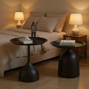 May include: Two black side tables with round tray tops and wide, bell-shaped bases. The left table holds a stack of books and a small abstract sculpture. The right table holds a stack of books with the word ARCHITECTURE printed on the spine.