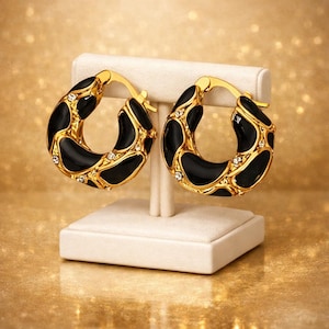 Può includere: Un paio di orecchini a cerchio color oro dal design spesso e massiccio, caratterizzati da inserti in smalto nero lucido e piccoli cristalli trasparenti incastonati. Gli orecchini sono esposti su un supporto chiaro, mostrando un'estetica metallica.