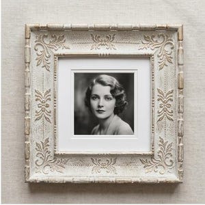 Op de afbeelding: Een vierkante fotolijst met een verweerde witte en gouden afwerking en gedetailleerde bloemmotieven in reliëf. De lijst bevat een wit passe-partout rondom een zwart-wit portretfoto van een persoon met golvend haar.