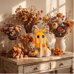May include: A white ceramic ghost figurine with a large bow on its head, illuminated from within, holding a small orange jack-o'-lantern bucket. The ghost features a patchwork pattern and is surrounded by several bouquets of dried autumn flowers.
