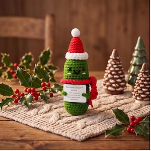 May include: A crocheted green pickle figurine wearing a red and white Santa hat and a red scarf. It holds a white sign that reads: Emotional Support Pickle. I will always be around to let you know that you are a BIG DILL!