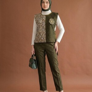 May include: A sleeveless vest featuring a split design with a brown patterned side and a dark olive green ribbed side, worn over a white long-sleeve shirt. The outfit includes dark olive green trousers, a dark olive green hijab, and a small handbag.