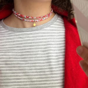 Peut inclure: Un collier ras-du-cou en tissu à double rang orné d'un motif floral rouge et blanc. Le collier comporte une petite breloque en forme d'étoile dorée suspendue au rang inférieur, reposant contre le cou.