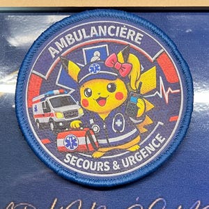May include: A circular embroidered patch featuring a yellow character wearing a blue uniform, a cap with a medical star, and a pink bow. The patch includes a white ambulance, a red medical bag, a radio, and the text AMBULANCIÈRE SECOURS & URGENCE.
