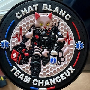 Peut inclure: Un écusson tactique circulaire noir représentant un chat blanc habillé en secouriste. Le chat porte un gilet noir avec l'inscription MEDIC, un sac médical rouge et une bouteille blanche. Le texte CHAT BLANC et TEAM CHANCEUX entoure le chat.