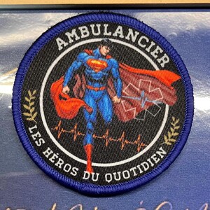 May include: A circular embroidered patch with a blue border featuring a graphic of a superhero in a blue suit and red cape. The text AMBULANCIER and LES HÉROS DU QUOTIDIEN surrounds the figure, along with a red medical star and an orange heartbeat line.
