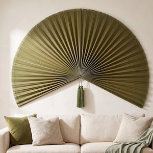 May include: A large, semi-circular wall fan in a deep olive green color, featuring fine vertical pleats radiating from a central point. A matching olive green tassel hangs from the center of the fan, which is mounted on a plain wall.