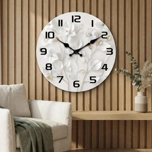 May include: A circular wall clock featuring a white background with a subtle, embossed-style floral and leaf pattern in shades of white and gold. The clock has bold black Arabic numerals and two black metal hands indicating the time.