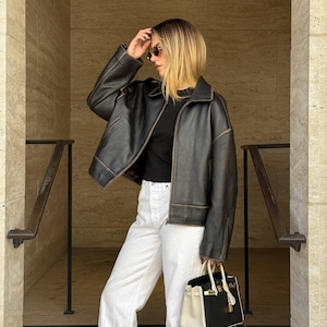May include: A black leather jacket with a collar and long sleeves, worn over a black top and white denim pants. A two-toned black and white handbag with a gold-tone lock and handle is held at the side. The jacket features visible stitching details.