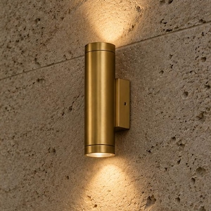 Può includere: Una lampada da parete cilindrica con finitura in ottone spazzolato. L'apparecchio presenta due aperture, una superiore e una inferiore, che proiettano un fascio di luce caldo e focalizzato verso l'alto e verso il basso sulla parete in pietra.