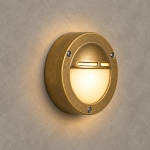May include: A circular wall-mounted light fixture with a weathered brass finish. The fixture features a semi-circular frosted glass lens that emits a warm glow, secured by three visible screws on the outer metal frame, creating a soft illumination.