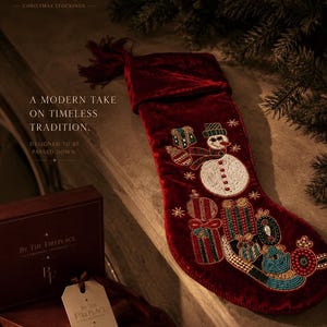 Puede incluir: Un calcetín navideño de terciopelo rojo intenso con abalorios detallados de un muñeco de nieve y regalos envueltos. El calcetín incluye una borla y viene con una caja a juego y una etiqueta que dice By The Fireplace Christmas Stockings.