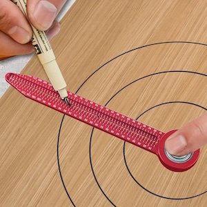 May include: A red metal circular marking tool featuring a long, flat arm with a series of small holes and printed numerical markings. The tool is positioned to draw concentric circles, with a pen tip inserted into one of the holes for precision.