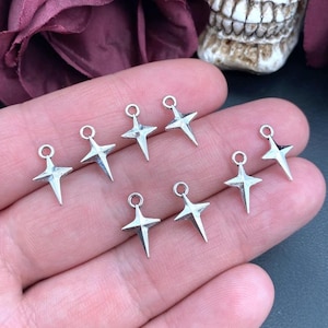Könnte beinhalten: Sieben kleine, silberfarbene Metallanhänger in Form von vierzackigen Sternen. Jeder Anhänger hat einen Mittelpunkt mit vier ausstrahlenden Strahlen und eine kleine kreisförmige Öse an der Oberseite zur Befestigung. Glatte, metallische Oberfläche.