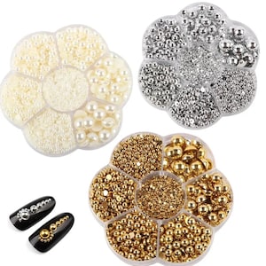 Può includere: Tre contenitori in plastica trasparente a forma di fiore riempiti con mezze perle bianche, argentate e dorate di varie dimensioni. Sono mostrate due punte di unghie nere, ciascuna decorata con un motivo di perline metalliche argentate e dorate.