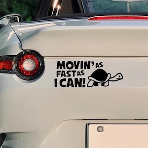 Könnte beinhalten: Ein schwarzer Vinyl-Aufkleber auf der Rückseite eines weißen Autos. Der Aufkleber zeigt eine stilisierte schwarze Schildkröte und den Text "MOVIN' AS FAST AS I CAN!" in fetten, schwarzen, serifenlosen Großbuchstaben neben dem roten Rücklicht.