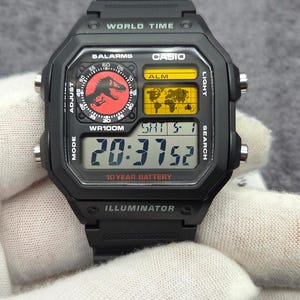 Può includere: Un orologio digitale nero con cinturino in resina e quadrante quadrato con la scritta WORLD TIME, CASIO, 5 ALARMS, WR100M, 10 YEAR BATTERY e ILLUMINATOR. Il display presenta un grafico di un dinosauro rosso, una mappa del mondo gialla e l'ora digitale.