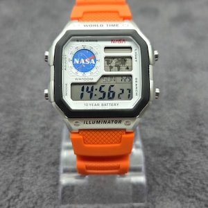 Op de afbeelding: Een digitaal polshorloge met een feloranje kunsthars band en een zilverkleurige rechthoekige kast. De wijzerplaat toont het NASA-logo, een wereldkaart en de tekst "WORLD TIME", "5 ALARMS", "WR100M", "10 YEAR BATTERY" en "ILLUMINATOR".