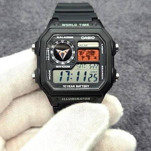 Op de afbeelding: Een zwart digitaal polshorloge met een vierkante kast, een zwarte kunststof band en een multifunctioneel display. De wijzerplaat bevat een wereldkaart, een ronde analoge sub-wijzerplaat en de tekst WORLD TIME, CASIO, 5 ALARMS, WR100M en 10 YEAR BATTERY.