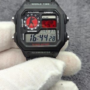 Puede incluir: Un reloj digital Casio negro con caja cuadrada y correa de resina negra. La esfera presenta un mapa mundial, una subesfera circular de estilo analógico con una silueta y una pantalla digital que muestra la hora 16:44:28.