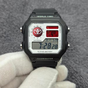 Pode incluir: Um relógio de pulso digital preto com caixa quadrada, bracelete de resina preta e mostrador branco. O visor mostra o logótipo da Toyota, um mapa-múndi e a hora 7:28. O texto inclui WORLD TIME, 5 ALARMS, WR100M, 10 YEAR BATTERY e ILLUMINATOR.
