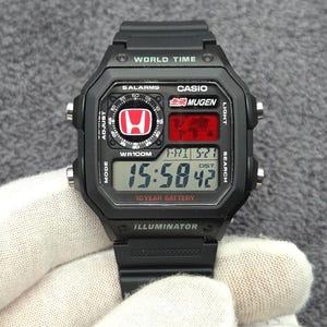Pode incluir: Um relógio digital Casio preto com caixa quadrada, pulseira de resina e exibição de hora mundial. O mostrador inclui um logótipo Honda vermelho, a marca Mugen, um relógio digital marcando 15:58:42, um mapa mundial e bateria de 10 anos.