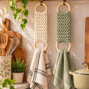 Op de afbeelding: Twee hangende keukendoekhouders met crème en saliegroene macramébanden bevestigd aan ronde houten ringen. Elke houder ondersteunt een wafeldoek, één met een crème en zwart ruitpatroon en de andere in effen saliegroen.