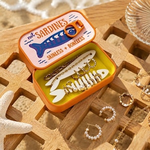 以下が含まれることがあります： 「not SARDINES」および「SKINLESS + BONLEESS」という文字と青い魚のグラフィックが描かれた蓋付きのオレンジ色の金属製缶。中には、魚の形をした2つの白いジュエリーホルダーがあり、パール付きのものを含むゴールドトーンのフープピアスが飾られています。