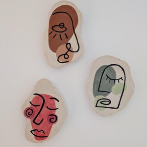Peut inclure: Trois plaques murales en céramique blanc cassé de forme irrégulière présentant des visages abstraits. Chaque plaque arbore un dessin au trait noir unique superposé à des formes peintes en terre cuite, vert sauge et rouge sourd.