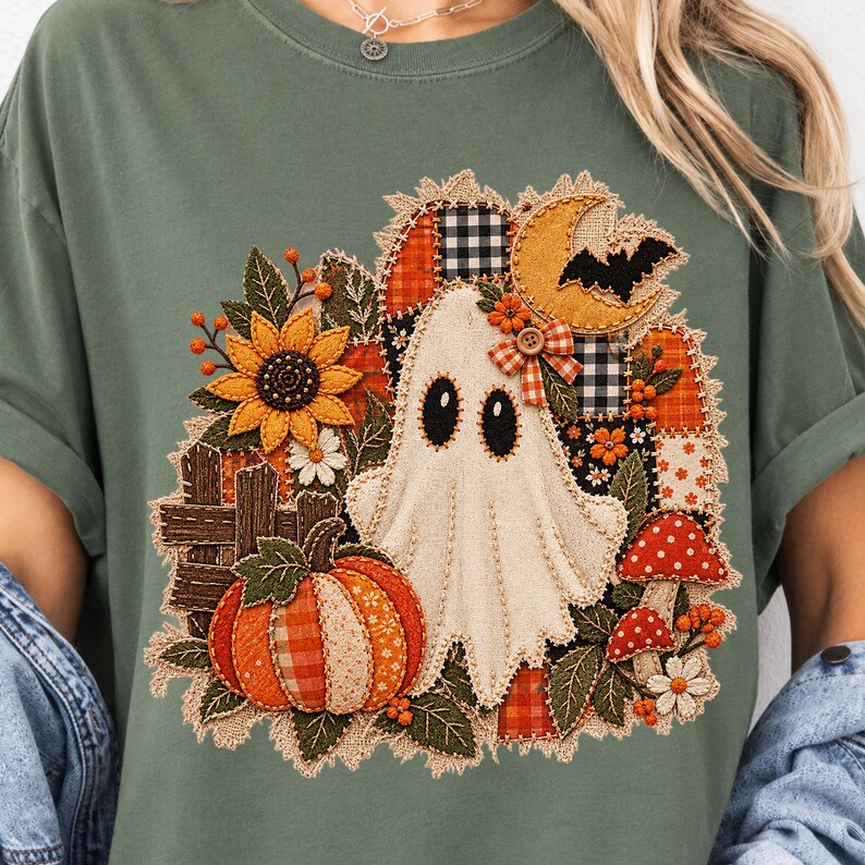 Peut inclure: Un t-shirt vert sauge avec un motif central repr&eacute;sentant un fant&ocirc;me blanc entour&eacute; d'un design style patchwork. L'illustration comprend une citrouille, un tournesol, un croissant de lune, une chauve-souris, des champignons et une cl&ocirc;ture en bois.