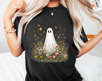 Peut inclure: Un t-shirt noir arborant un motif central représentant un fantôme blanc debout dans une prairie fleurie, remplie de fleurs sauvages colorées, de feuillage vert et de petits papillons orange. Le design inclut des fleurs roses, jaunes et blanches.