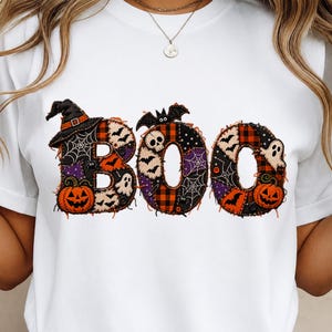 Op de afbeelding: Een wit t-shirt met het woord BOO in grote letters. De letters zijn gevuld met een patchwork van Halloween-patronen, waaronder oranje pompoenen, zwarte vleermuizen, witte geesten, spinnenwebben en rood-zwarte ruitjesstoffen.