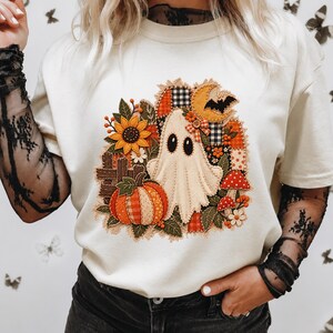 Peut inclure: Un t-shirt couleur cr&egrave;me arborant un motif central compos&eacute; d'un fant&ocirc;me, d'une citrouille, d'un tournesol, de champignons et d'un croissant de lune. Le design pr&eacute;sente une esth&eacute;tique patchwork avec divers motifs de tissus et textures.