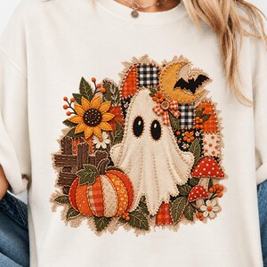 Peut inclure: Un t-shirt couleur cr&egrave;me arborant un motif central compos&eacute; d'un fant&ocirc;me blanc, d'une citrouille, d'un tournesol, d'un croissant de lune, d'une chauve-souris et de champignons rouges. Le design inclut des textures patchwork et des boutons.
